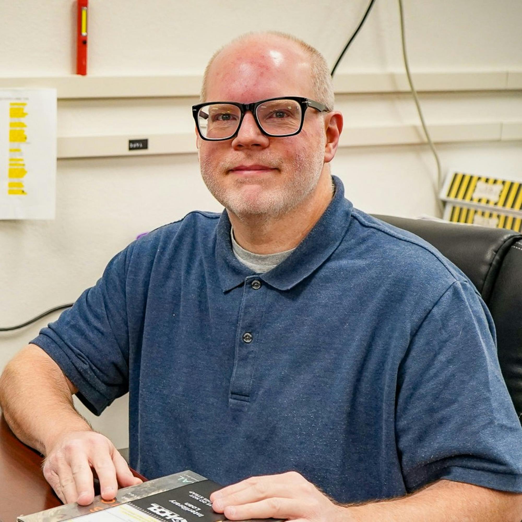 Home Bald man wearing glasses sitting at a desk in office environment holding a book.