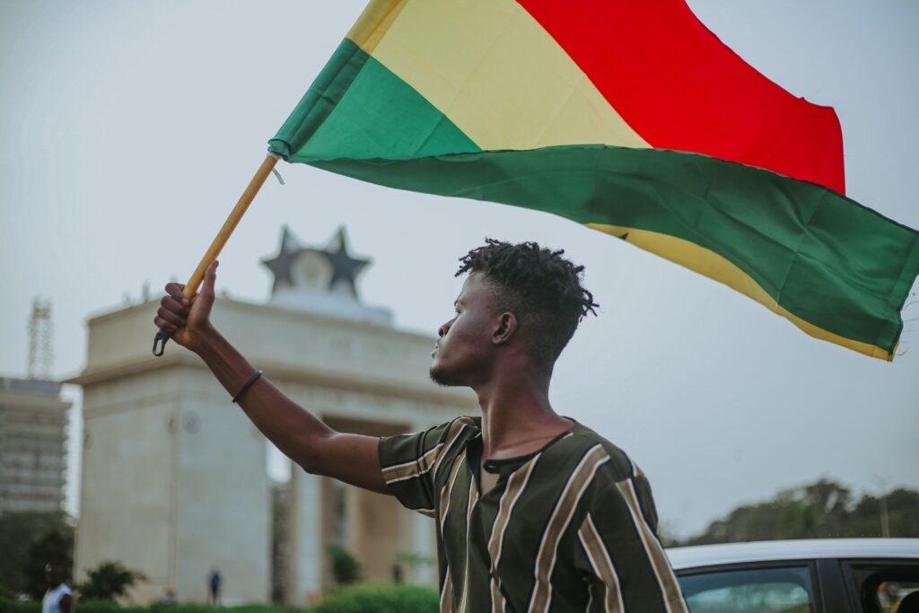 About Us African male with dreadlocks raising flag of Ghana country with colorful stripes while looking away in town